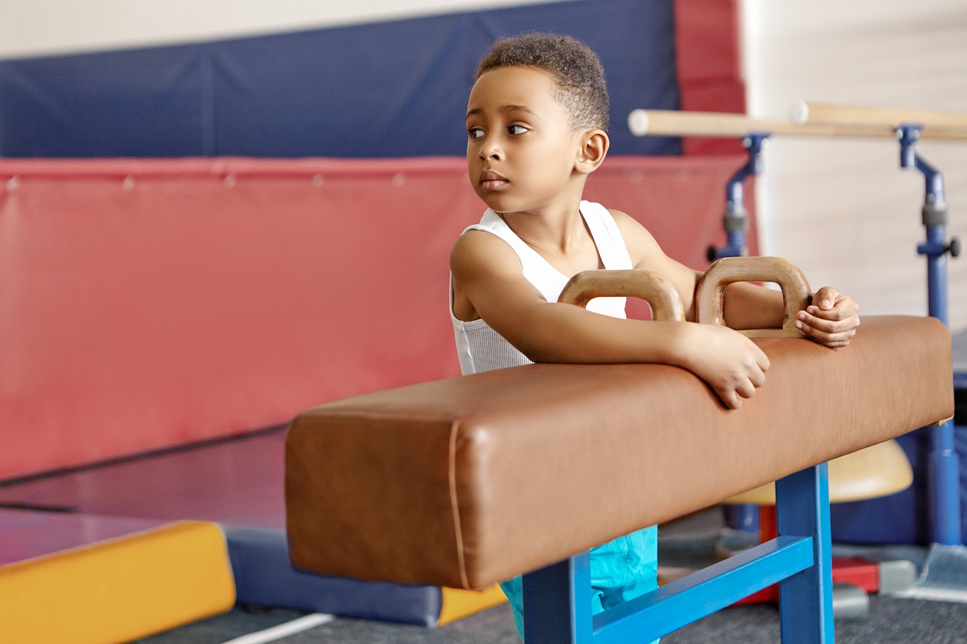 Cute handsome dark skinned schoolboy exercising on pommel horse with brown leather cover. Portrait of African child using artistic gymnastics apparatus at gym. Childhood, activity and sports