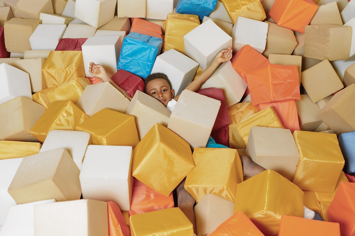 Happy chilldhood, entertainment and modern active lifestyle concept. Adorable active African male child lying among colorful soft fabric cubes while spending weekend in playroom at city mall